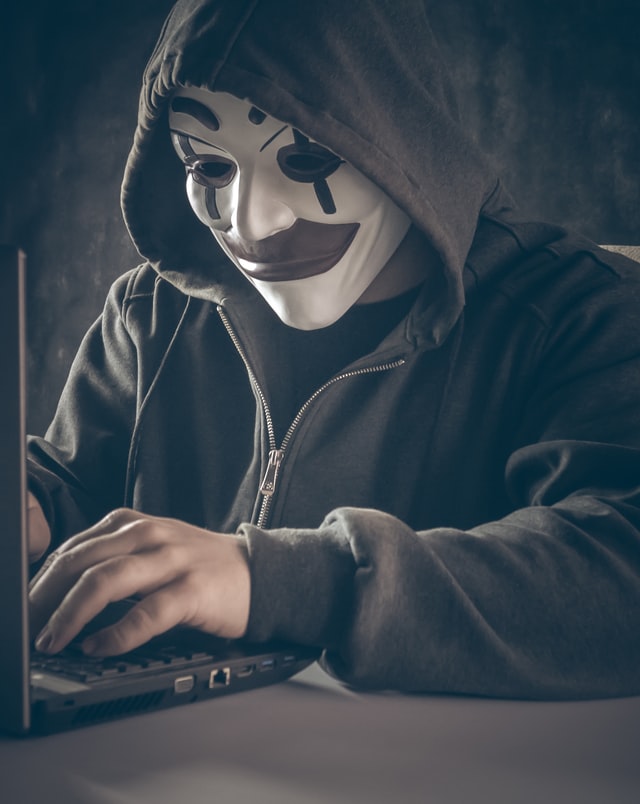 A masked man in a hoodie types away on his laptop. A hacker.
