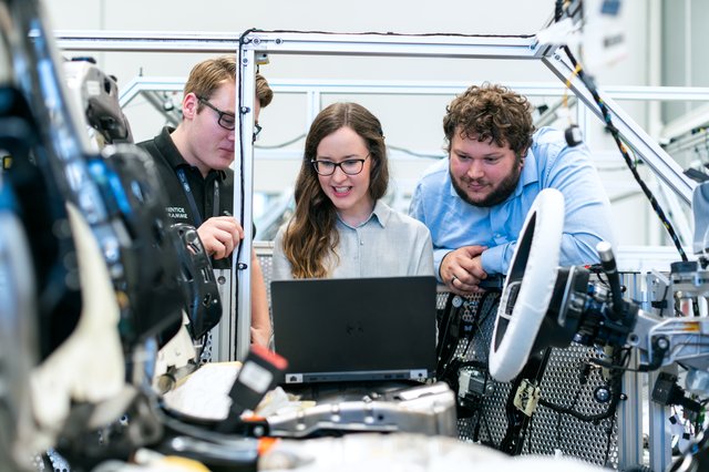 3 people looking at a laptop inside a car manufacturing facility